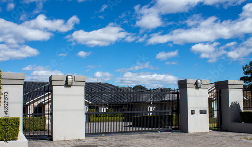 Bild auf Leinwand Black metal driveway entrance gates set in brick fence with private house in bac