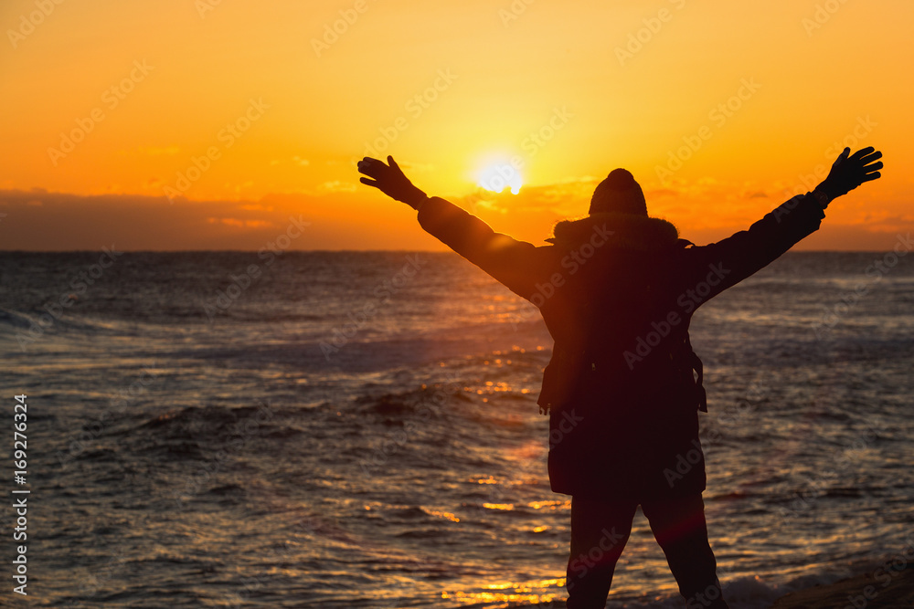 A traveler with a backpack looks at the sunrise in the Japanese sea and raises his hands up.