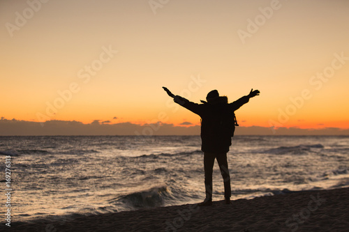 A traveler with a backpack looks at the sunrise in the Japanese sea and raises his hands up.