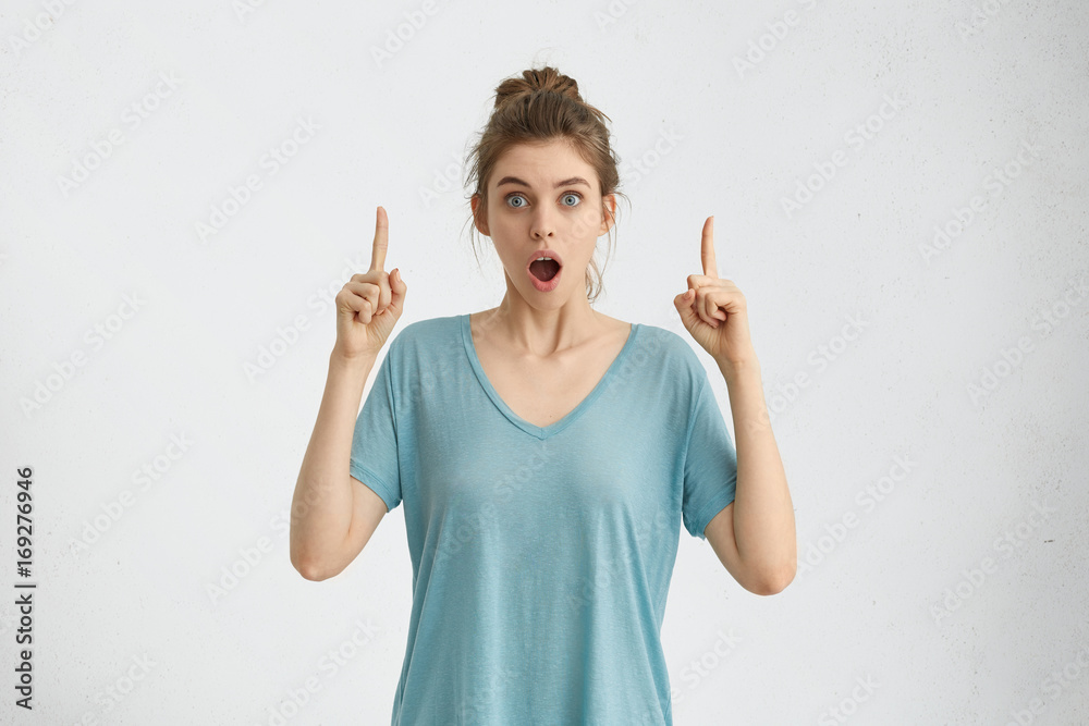 Astonished amazed teenage girl with hair knot looking at camera with mouth dropped open and eyes popped out, pointing fingers up, indicating something surprising and shocking on white studio wall Astonished amazed teenage girl with hair knot looking at camera with mouth dropped open and eyes popped out, pointing fingers up, indicating something surprising and shocking on white studio wall