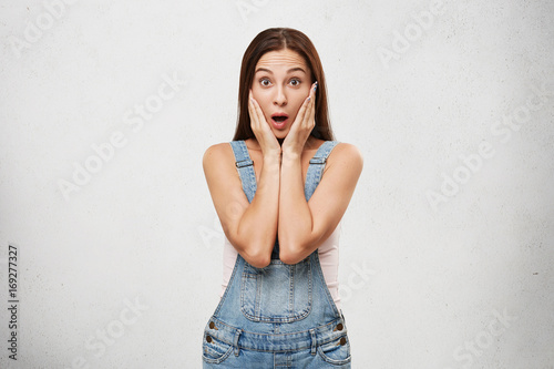 Portrait of astonished emotional brunette girl in stylish jeans jumpsuit opening mouth and holding face with both hands, shocked with unexpected news, her look and gestures expressing surprise