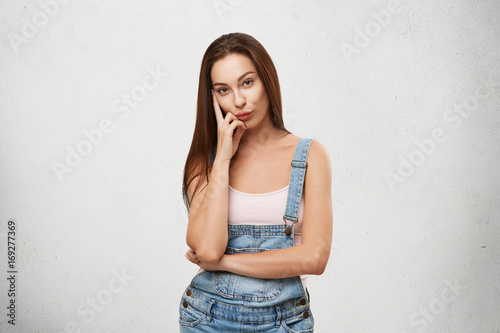 Let me think. Smart cunning quickwitted beautiful gir in jeans jumpsuit looking pensive while plotting or scheming something, holding hand on her cheek. Human facial expressions, reaction and attitude photo