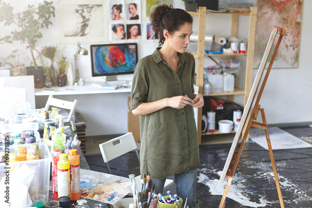 Art and inspiration. Indoor shot of hesitant young female artist wearing  jeans and khaki shirt standing in spacious workshop interior in front of  easel, evaluating painting that she just finished Stock Photo