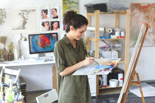 Indoor portrait of happy excited young woman artist in shirt of military color holding palette and paintbrush while working on painting in her workshop, standing in front of easel and smiling