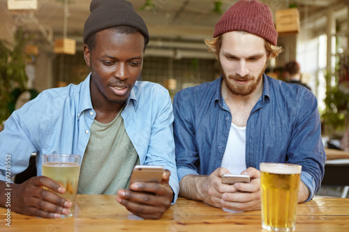 Technologies  online communication and internet addiction. Handsome bearded Caucasian guy and his African American friend or partner enjoying fresh beer at pub and browsing social networks on mobiles