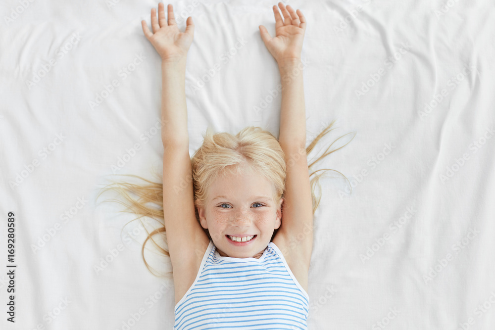 Adorable little girl with blonde hair and freckles, waking up in morning, stretching on white bedclothes, having pleasant smile after good sleep. Small kid smiling into camera while lying in bed Adorable little girl with blonde hair and freckles, waking up in morning, stretching on white bedclothes, having pleasant smile after good sleep. Small kid smiling into camera while lying in bed