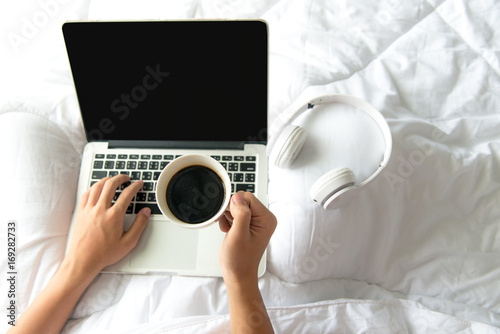 Male sitting on white bed surfing internet or working on laptop and listening to musical media online with wireless headphone and drink coffee in morning relax mood. Music and Lifestyle Concept.