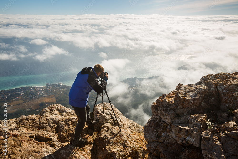 Man standing with a tripod and camera on a high mountain peak above clouds, city and sea. Professional photographer adjusting dslr settings on rocky summit.
