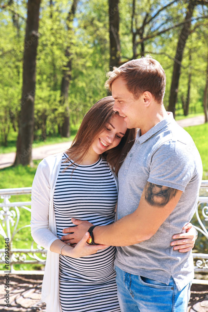 Fototapeta premium Handsome man and his beautiful pregnant wife are hugging and smiling while standing in park