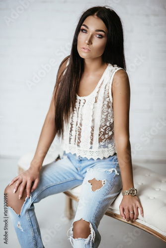 Young beautiful showy girl with luxurious, shiny hair sitting on a bar stool. Brown hair, blue eyes, big lips. Blue ragged jeans. Fashion, beauty. Salon. photo