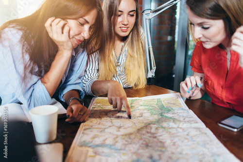 Three girlfriends planning their vacation sitting at table around map choosing the destination