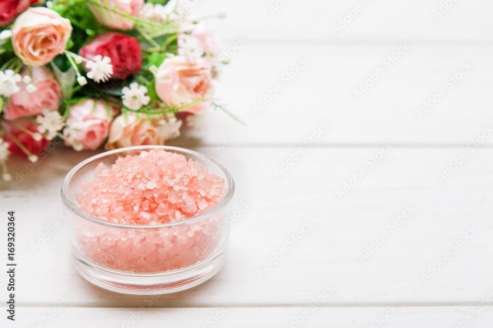 Red sea salt in a small glass bowl and roses bouquet behind on a white wooden table, text space template Red sea salt in a small glass bowl and roses bouquet behind on a white wooden table, text space template