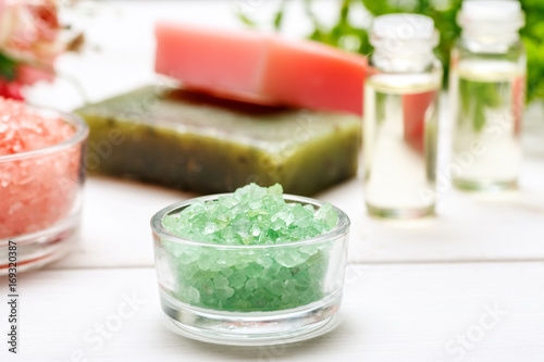 Green sea salt in a small glass bowl, handmade soap and few small bottles with liquid inside behind on a white wooden table