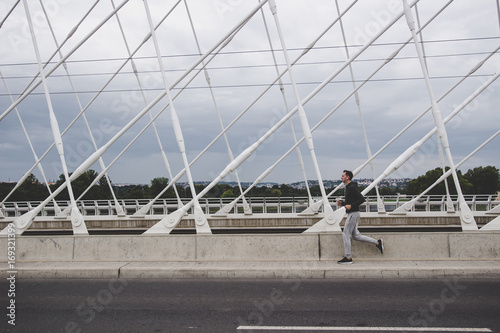 Young man running on the modern bridge in the city, listening music on smartphone