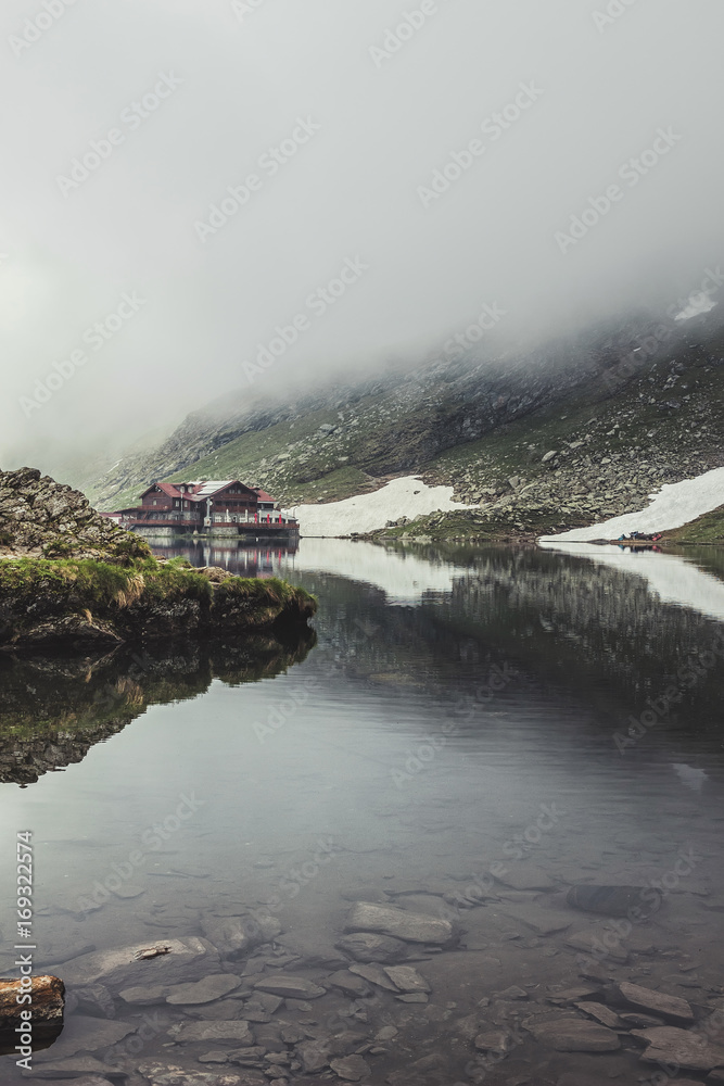 Nature landscape of amazing glacier lake Balea with house on shore and Fagaras mountains with white strips of snow reflected in the water at Carpathians, Romania