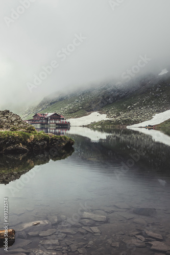 Nature landscape of amazing glacier lake Balea with house on shore and Fagaras mountains with white strips of snow reflected in the water at Carpathians, Romania