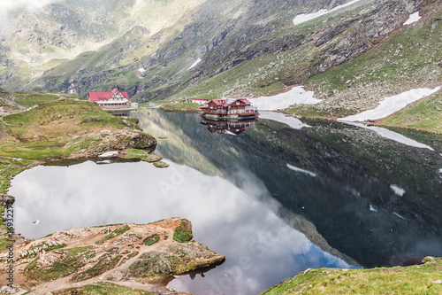 Nature landscape of amazing glacier lake Balea with house on shore and Fagaras mountains with white strips of snow reflected in the water at Carpathians  Romania