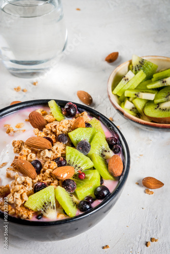Healthy diet breakfast: smoothies bowl, with yogurt, fresh blueberries (black currant), kiwi, granola oatmeal, almond nuts. On a white stone table, with a glass of water and ingredients. Copy space