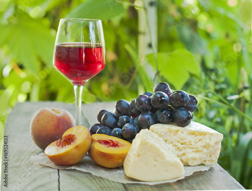 Cheese with a glass of red wine and a brush of grapes on a wooden table, in a garden with a vine