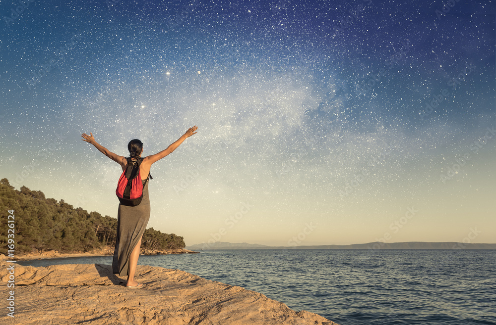 A young woman on the seashore raises her hands to the sky against the starry sky.