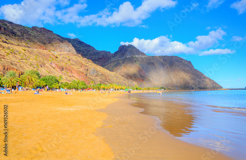 beach on the island of Tenerife in bright sunny day, a great number of vacationers ashore, on a background mountains tower
