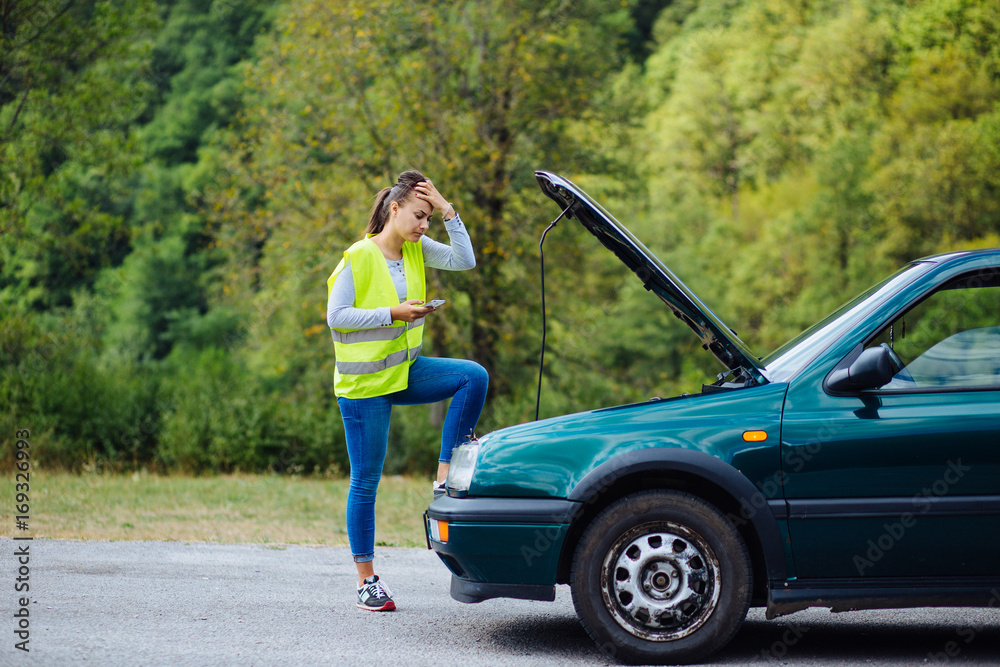 Worried young white female holding her head staring at the car engine of her broken car waiting for repair service Worried young white female holding her head staring at the car engine of her broken car waiting for repair service