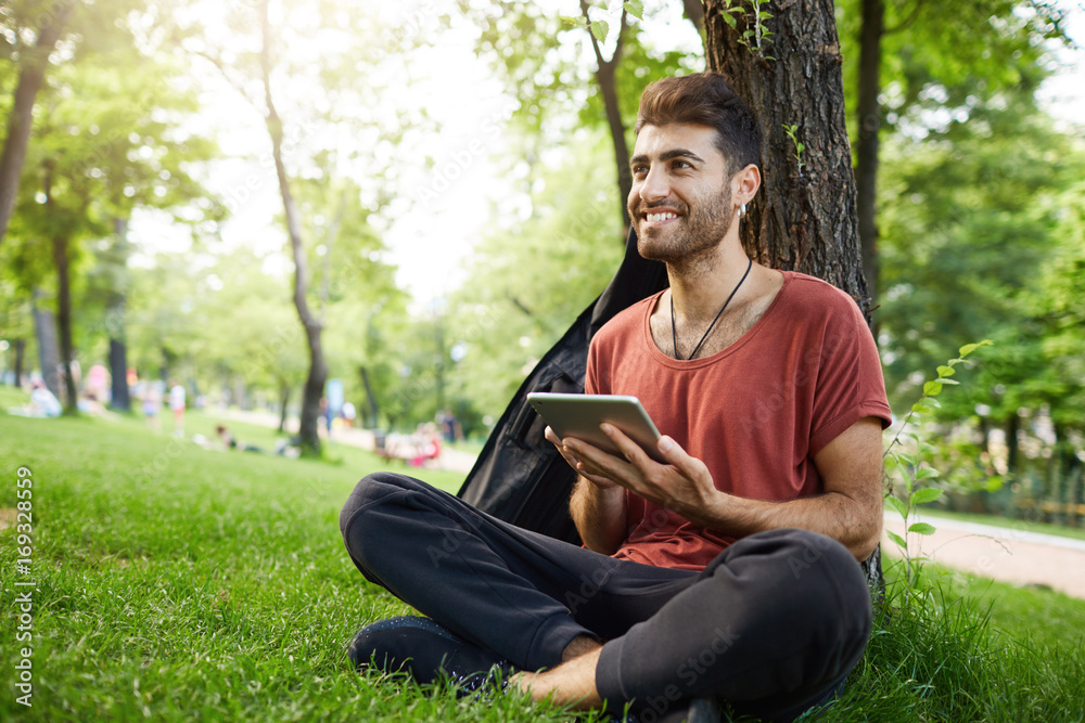 Attractive bearded male holding tablet in his hands. Good-looking smiling casually weared boy sitting cross-legged at park, using tablet. Attractive bearded male holding tablet in his hands. Good-looking smiling casually weared boy sitting cross-legged at park, using tablet.