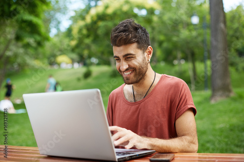 People, beauty and lifestyle concept. Shot of handsome man with beard and wide smile dressed in red T-shirt smiling broadly being happy to receive e-mail with good news, typing reply to this e-mail