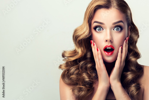 Woman with ren lips and nails surprise holds cheeks by hand .Beautiful girl  with curly hair surprised and shocked looks on you . Presenting your product. Expressive facial expressions