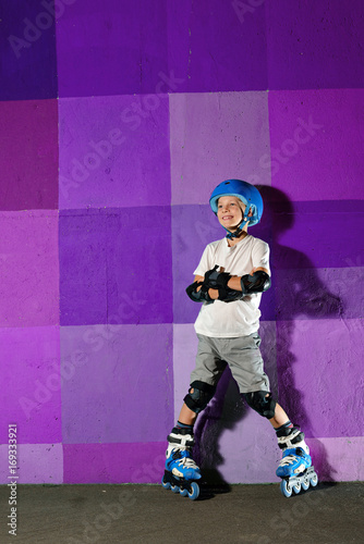 Cute little athletic boy on roller standing against the purple graffiti wall