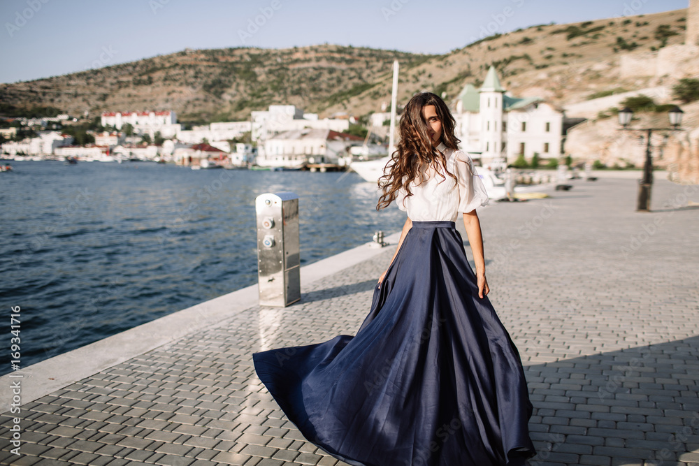 Beautiful girl walking on the pier by the sea near the yacht, boats in the blue long dress Beautiful girl walking on the pier by the sea near the yacht, boats in the blue long dress