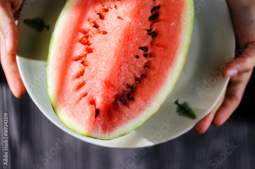 Slice of watermelon on white plate in woman hands ready for eating, top view with blurry black wood table background photo