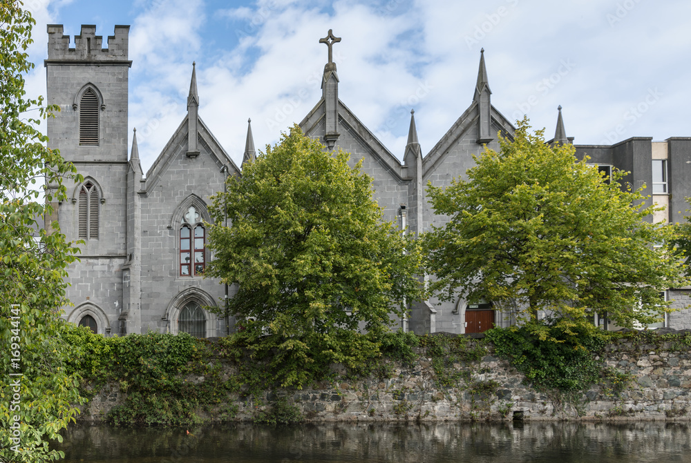 Naklejka premium Galway, Ireland - August 3, 2017: The gray stone Saint Vincent’s Convent of Mercy with its towers and crosses stands behind a couple of green trees along the Corrib River. Blue white skies.