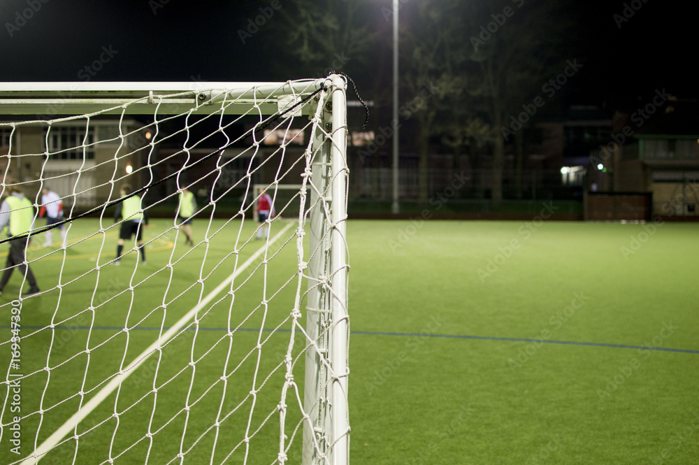 Fototapeta premium Behind Football Goalposts During Amateur Game On Floodlit Pitch