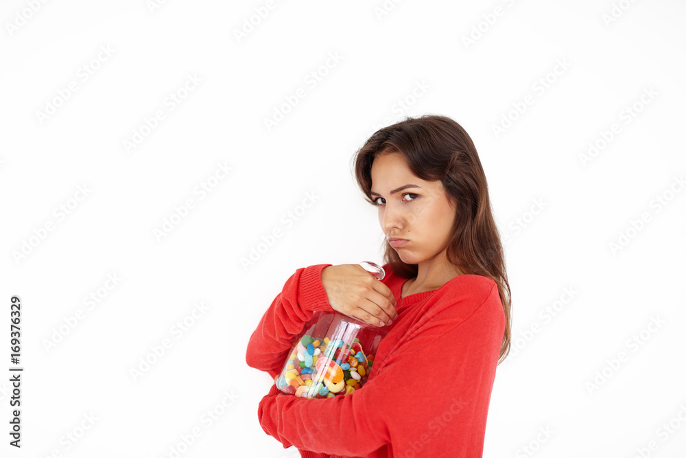 Beautiful greedy brown haired teenage girl of mixed race appearance holding tight big plastic jar with multicolored candies or jelly beans, about to eat everything by herself, not sharing with anyone Beautiful greedy brown haired teenage girl of mixed race appearance holding tight big plastic jar with multicolored candies or jelly beans, about to eat everything by herself, not sharing with anyone