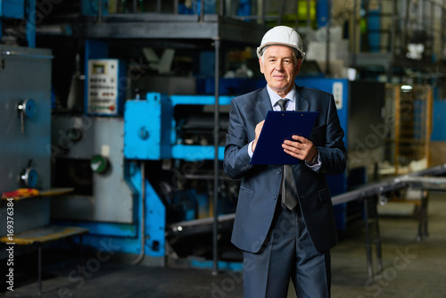 Portrait of smiling senior businessman wearing hardhat looking at camera holding clipboard in workshop of modern factory © Seventyfour
