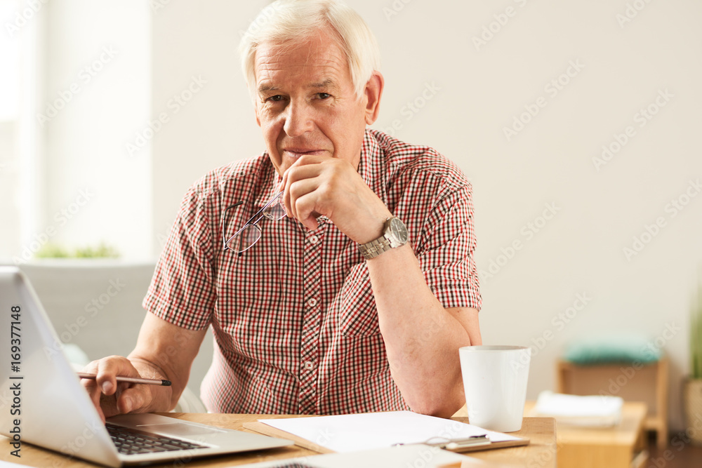 Portrait of modern senior man using laptop at home working and looking at camera, smiling Portrait of modern senior man using laptop at home working and looking at camera, smiling