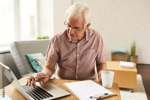 Portrait of senior man working with laptop at home counting finances and typing