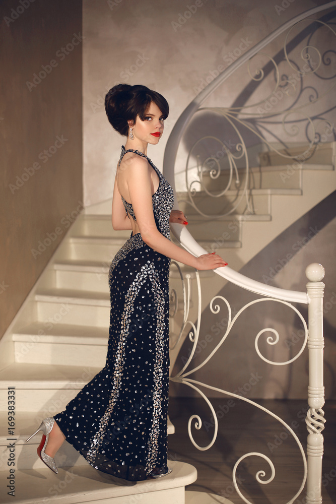 Beautiful Lady In Fashion Dress Posing On Front Staircase Elegant Brunette Sexy Woman In Long Gown Attractive Girl Model With Red Lips Makeup And Hairstyle In Modern Hall Interior Stock Photo