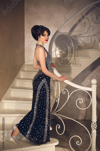 Beautiful Lady In Fashion Dress Posing On Front Staircase Elegant Brunette Sexy Woman In Long Gown Attractive Girl Model With Red Lips Makeup And Hairstyle In Modern Hall Interior Stock Photo