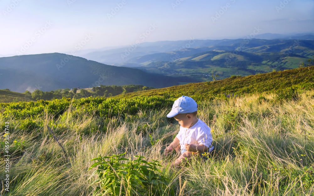 Little baby playing with grass in the mountains in summer. Beautiful mountains landscape in the background. Summer vacation in Ukrainian Carpathians for family with child. Little baby playing with grass in the mountains in summer. Beautiful mountains landscape in the background. Summer vacation in Ukrainian Carpathians for family with child.