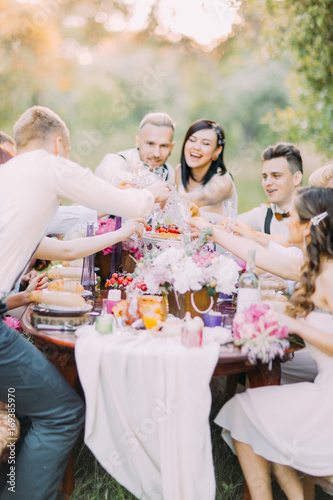 Happy close-up portrait of the delightful newlyweds and guests clinking their glasses and having fun at the setting table full of food and lovely flowers in the forest. photo