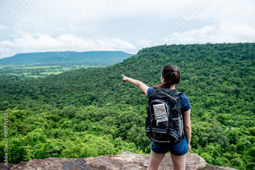 Young woman with backpack map behind bag standing on cliff's edge pointing to sky
