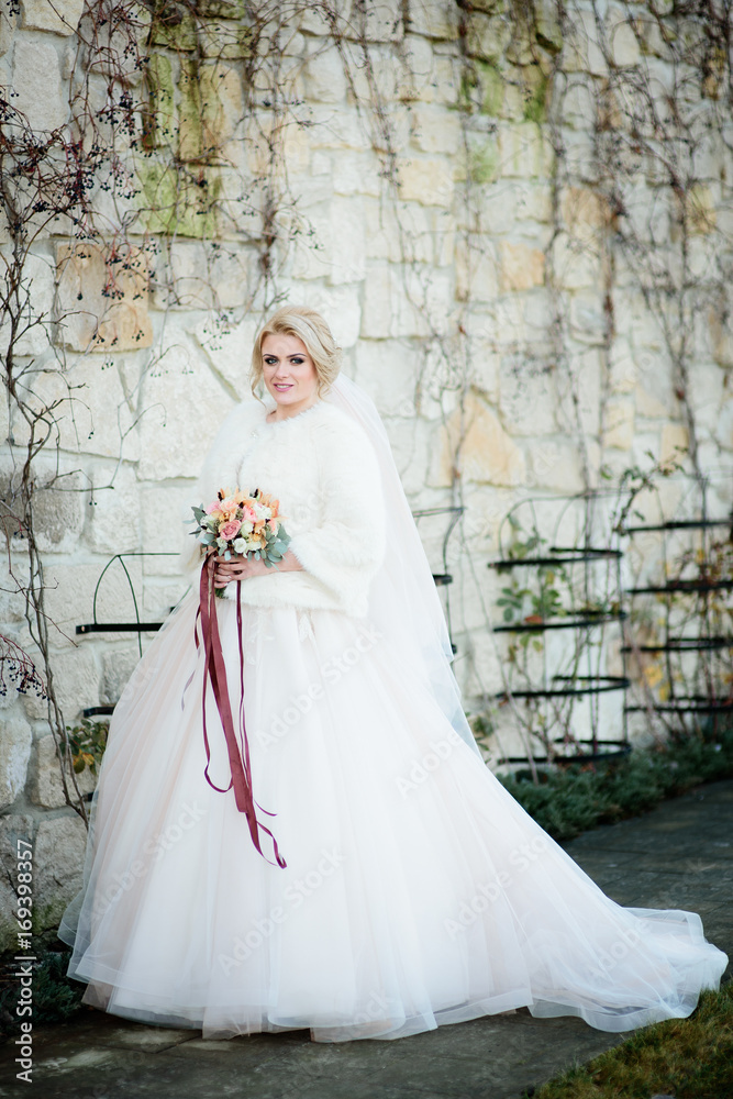 Obraz premium Magnificent bride in luxury dress and white coat stands before stone wall