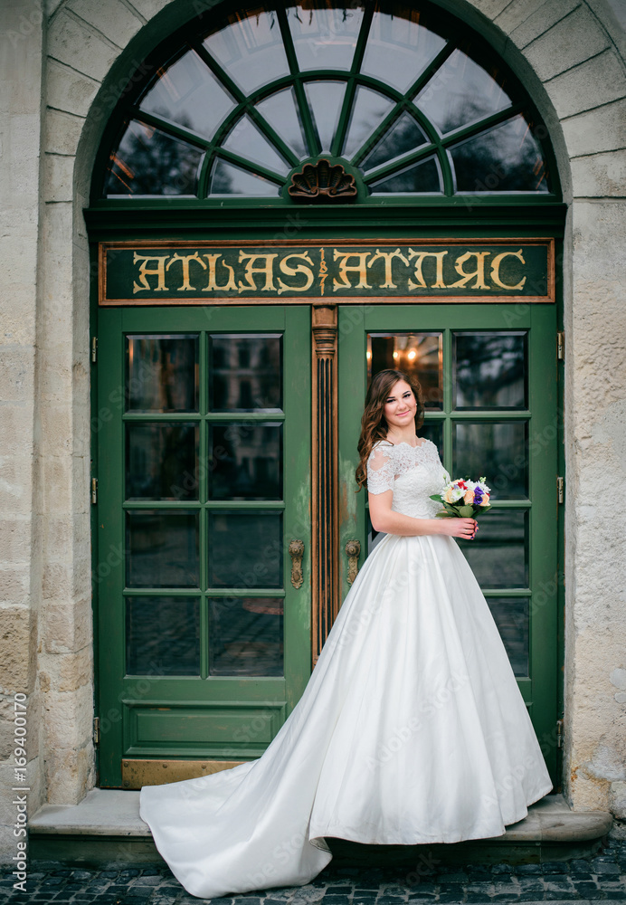 Obraz premium Curly bride in long dress stands before green door in white house