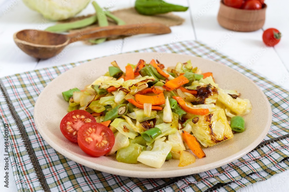 Vegetable stew - a mixture of baked cabbage, green beans, onions, carrots, cherry tomatoes, sweet pepper on a plate on a white wooden background. Vegan cuisine. Proper nutrition. Vegetable stew - a mixture of baked cabbage, green beans, onions, carrots, cherry tomatoes, sweet pepper on a plate on a white wooden background. Vegan cuisine. Proper nutrition.