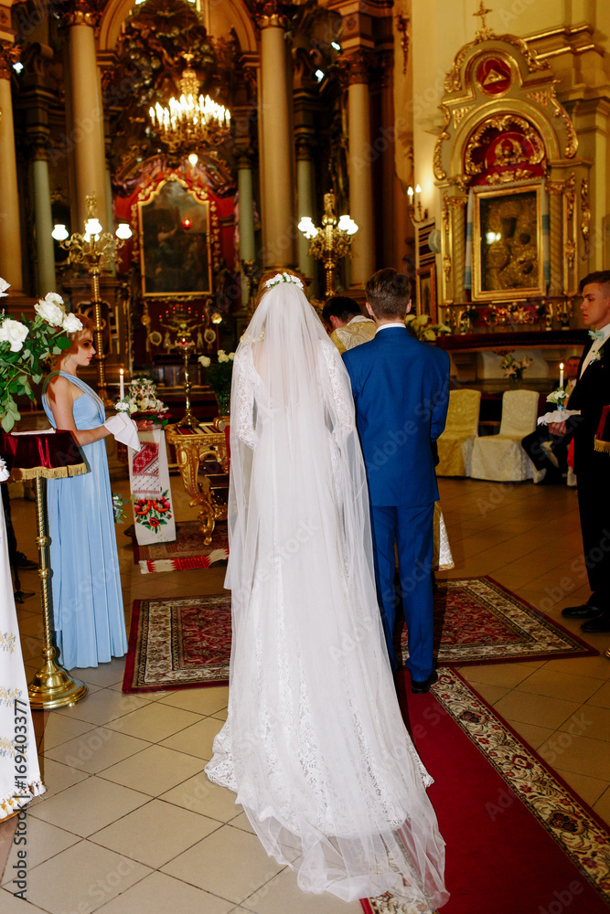 Look from behind at groom in blue suit and bride in long dress wtih long veil standing in church Look from behind at groom in blue suit and bride in long dress wtih long veil standing in church