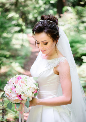 Tender shy bride poses with wedding bouquet in bright green forest