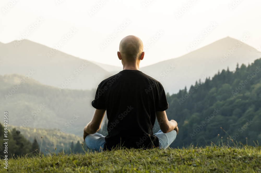 Skinny male person doing yoga sits in lotus position top of the hill in ukrainian carpathian mountains. Young man meditating in mountains at sunset 