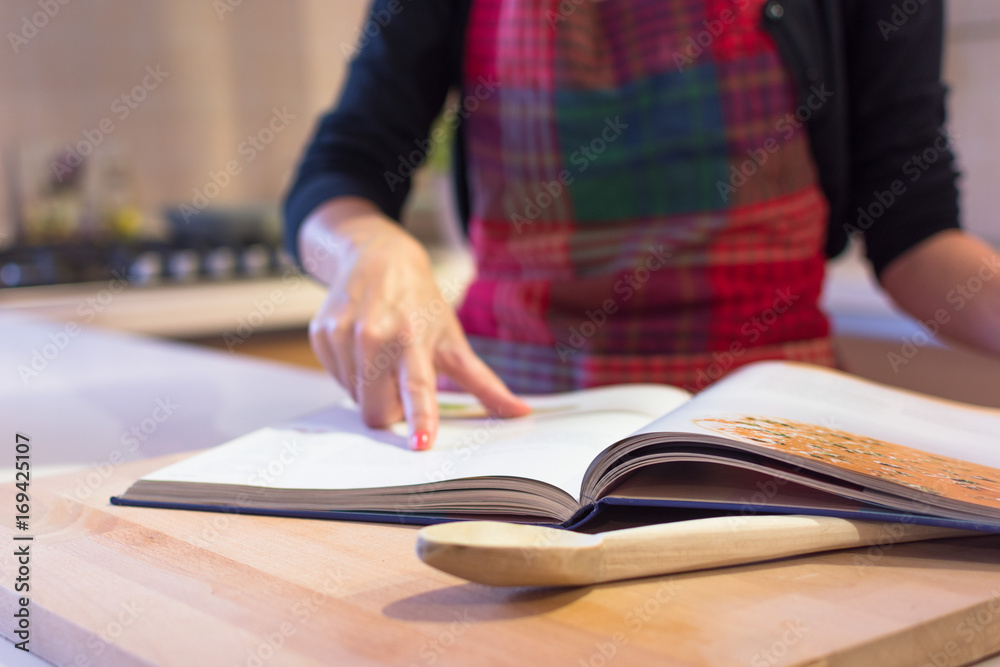 Close-up mid section of a woman, wearing a kitchen apron, reading a recipe book in the kitchen at home. Close-up mid section of a woman, wearing a kitchen apron, reading a recipe book in the kitchen at home.